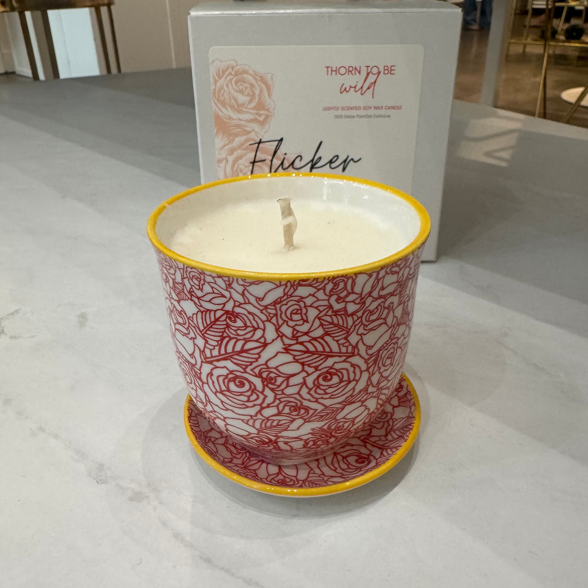 A candle in a white ceramic pot with a red rose pattern and yellow rim, sitting on a matching saucer in front of its white packaging box on a light marble countertop.