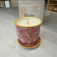 A candle in a white ceramic pot with a red rose pattern and yellow rim, sitting on a matching saucer in front of its white packaging box on a light marble countertop.