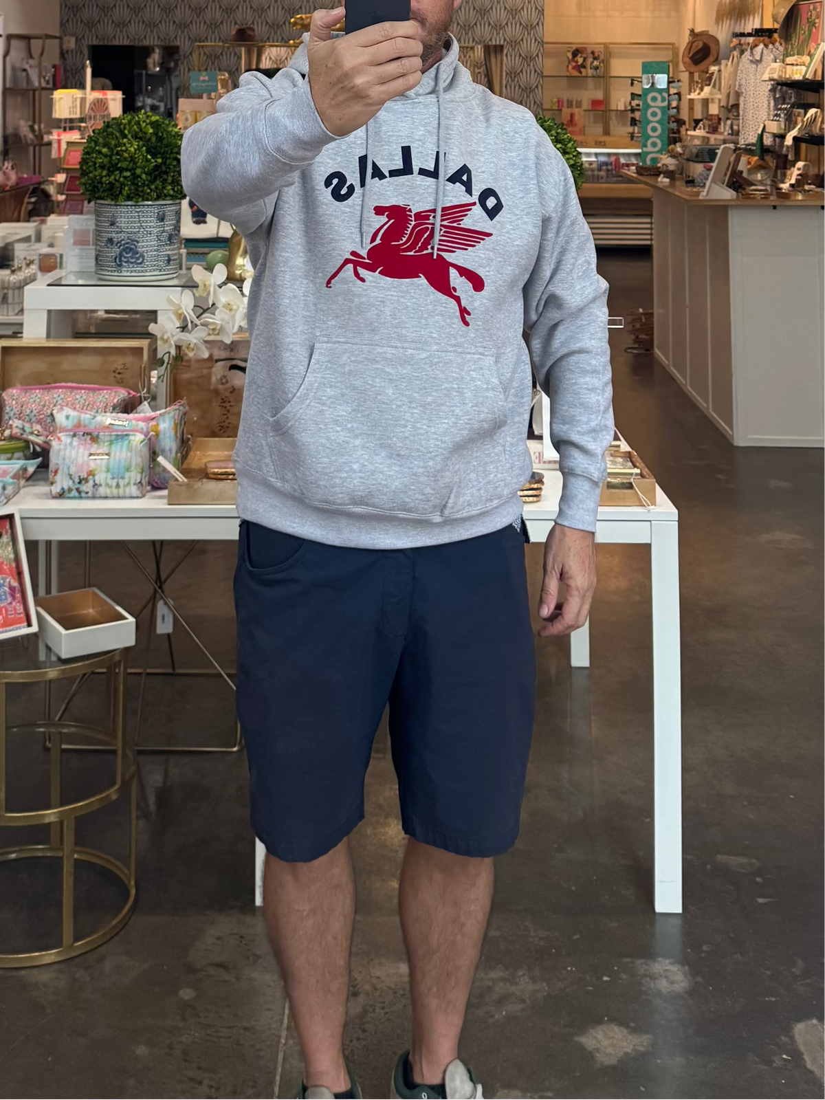 A man takes a mirror selfie inside a boutique shop, wearing a light gray Balenciaga hoodie with a red horse logo and navy blue shorts.