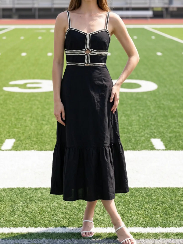 A woman stands on a football field wearing a black midi dress with a structured sweetheart bodice, silver rhinestone trim, and thin shoulder straps.