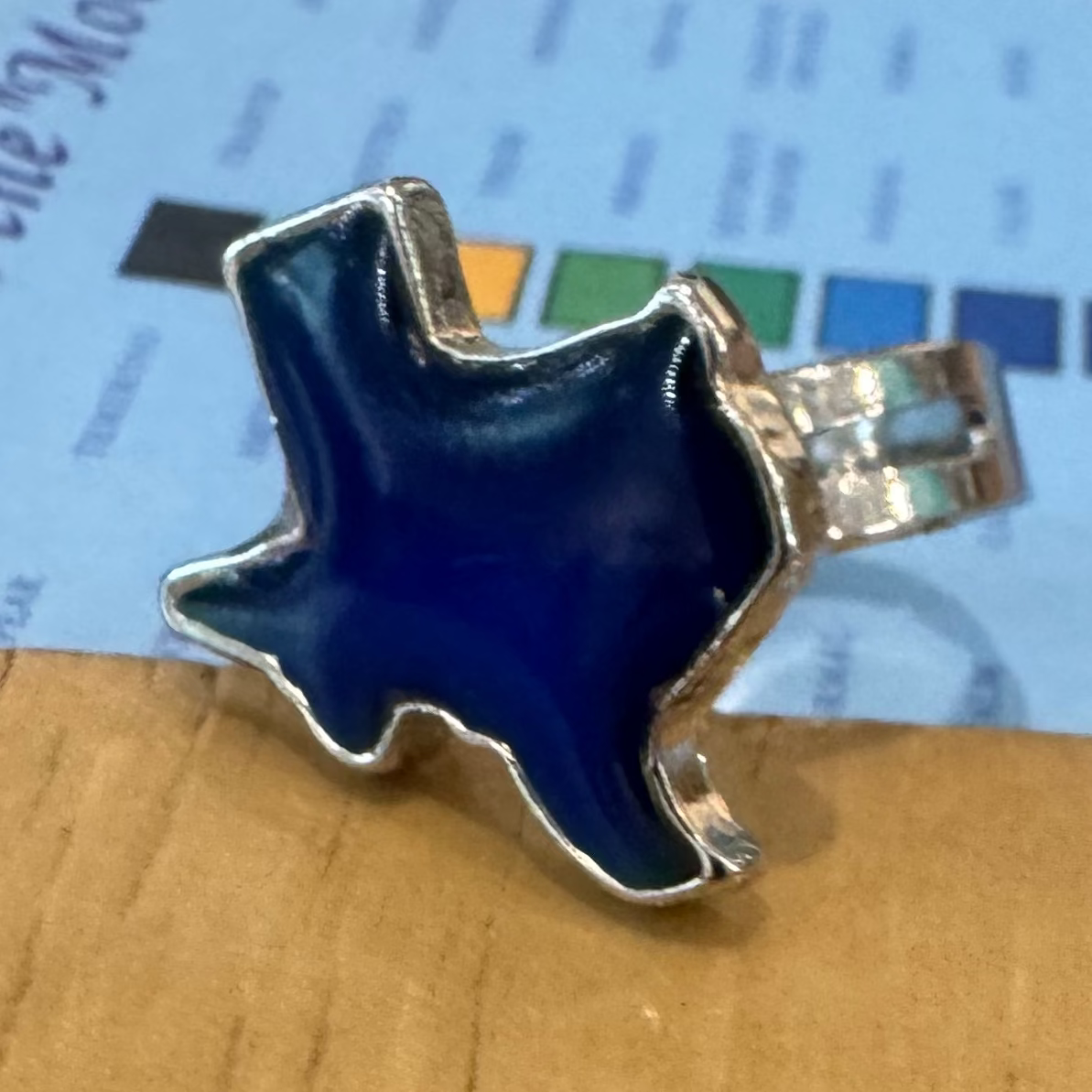 A silver-toned ring with a blue enamel star-shaped face, resting on a light blue color card over a wooden tabletop.