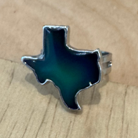 A silver ring shaped like the state of Texas, featuring a dark teal and green gradient enamel finish, resting on a light wood grain surface.